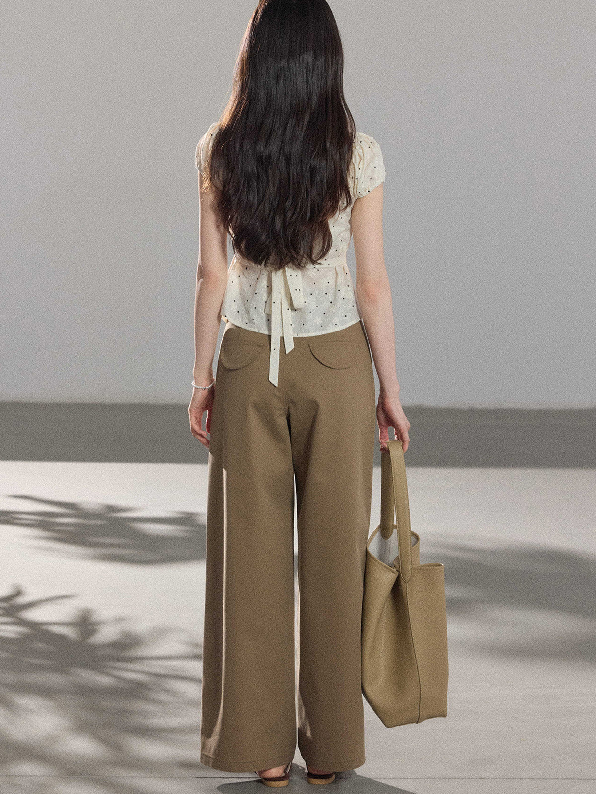 The back view of a woman wearing a cream polka dot top with side ties and brown wide-leg pants.