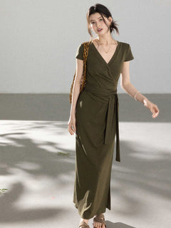 A woman in an olive green wrap dress with a V-neck and tie belt, walking with a woven bag.