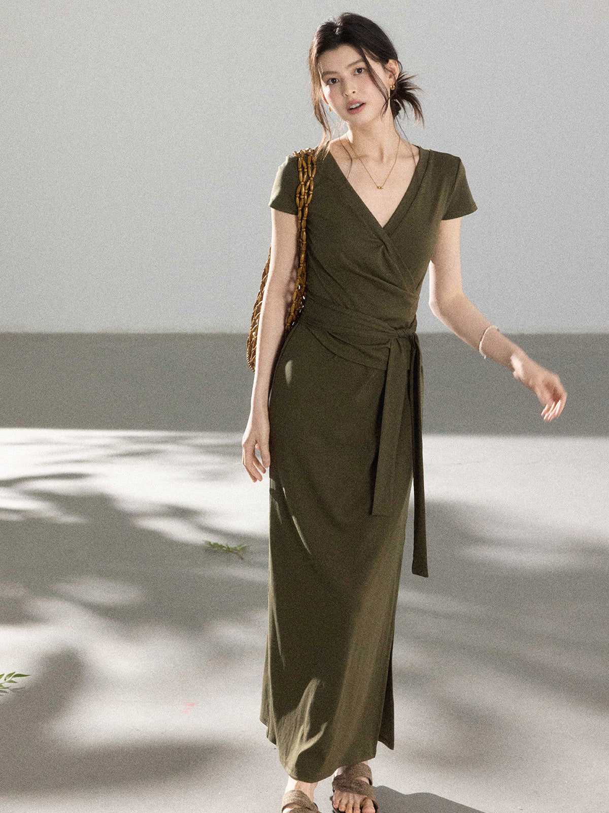 A woman in an olive green wrap dress with a V-neck and tie belt, walking with a woven bag.
