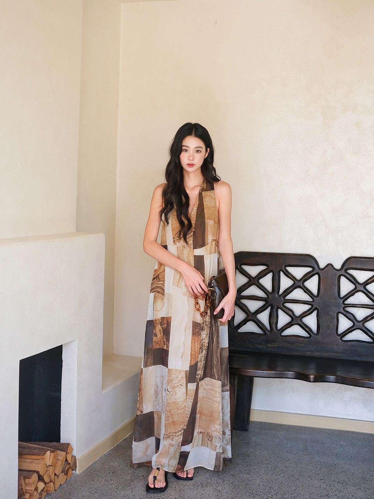 A woman standing in front of a wooden bench, wearing a long dress with a brown and beige patchwork pattern.