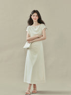 Cream off-shoulder dress featuring draped fabric at neckline and waist, side slit.