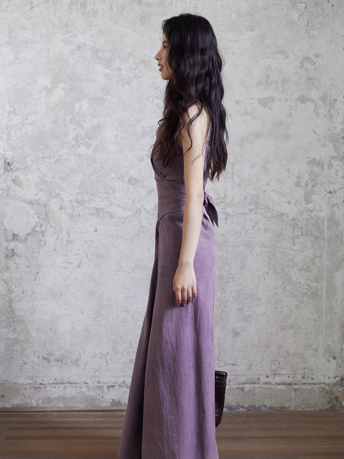 A side profile of a woman wearing a purple sleeveless maxi dress with a back tie belt.