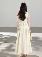 A woman in a cream sleeveless A-line floor-length dress with a back tie detail and pleated skirt.