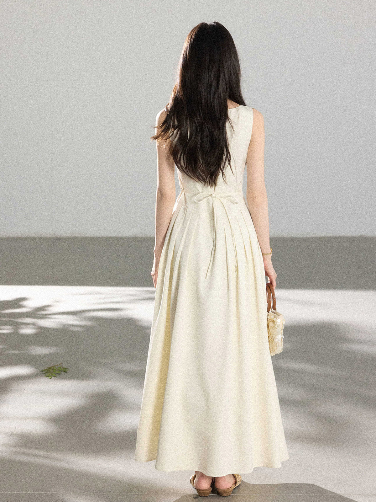 A woman in a cream sleeveless A-line floor-length dress with a back tie detail and pleated skirt.