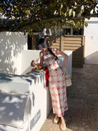 A woman in a long, sleeveless dress with a red diamond grid pattern and floral motifs stands by a white wall holding a beaded bag.