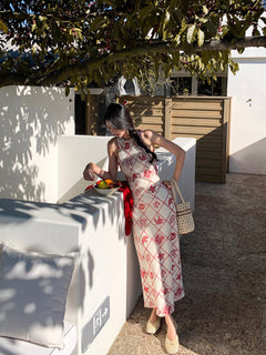 A woman in a long, sleeveless dress with a red diamond grid pattern and floral motifs stands by a white wall holding a beaded bag.
