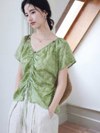 A woman wears a green tie-dye top with a ruched front and short sleeves, carrying a woven bag.