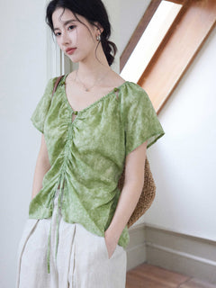 A woman wears a green tie-dye top with a ruched front and short sleeves, carrying a woven bag.