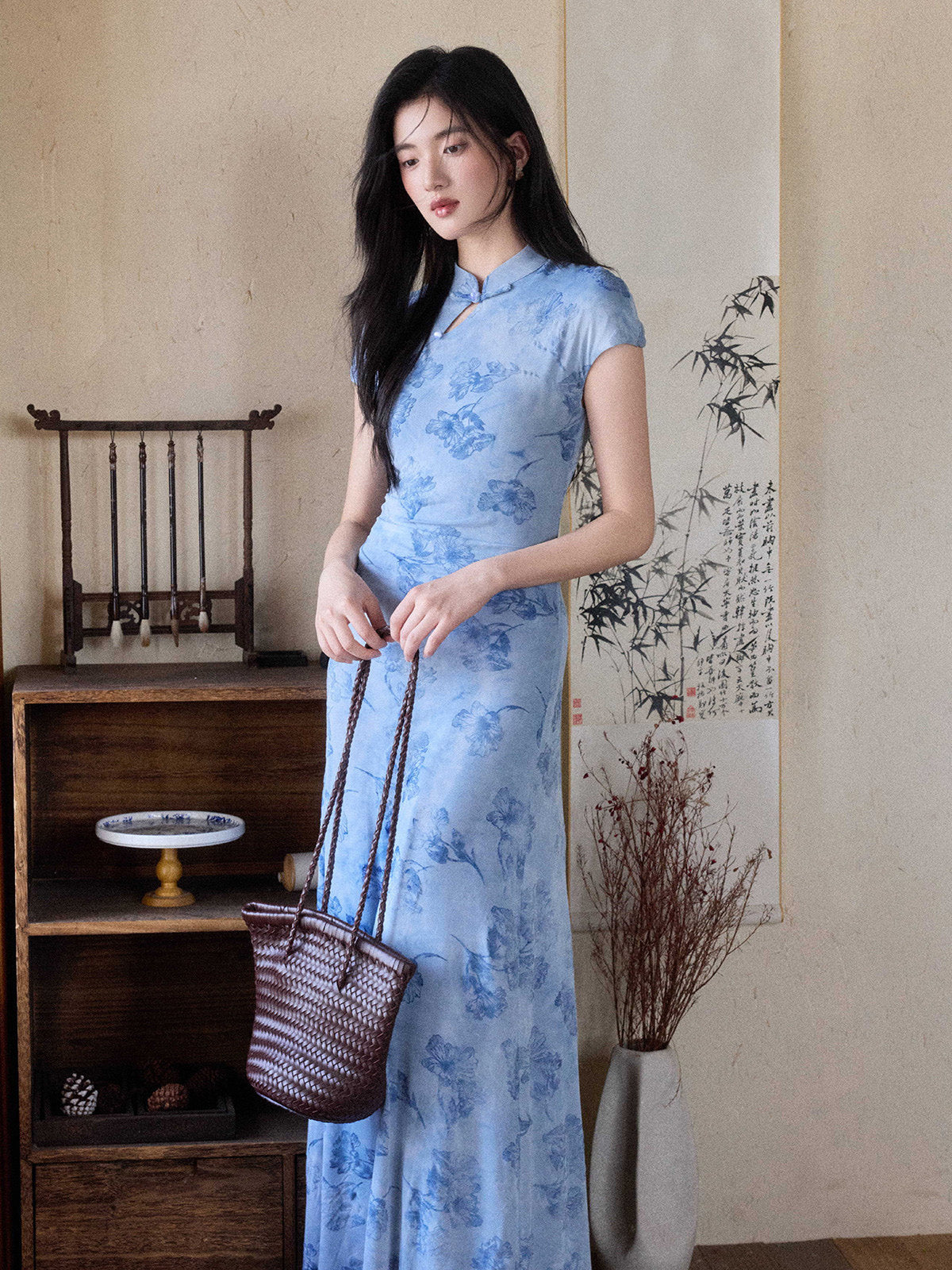 A woman in a light blue floral dress with a mandarin collar and keyhole opening, standing while holding a woven bag.