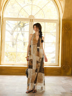 A woman standing by a large window, wearing a long dress with a brown and beige patchwork pattern.