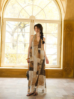 A woman standing by a large window, wearing a long dress with a brown and beige patchwork pattern.