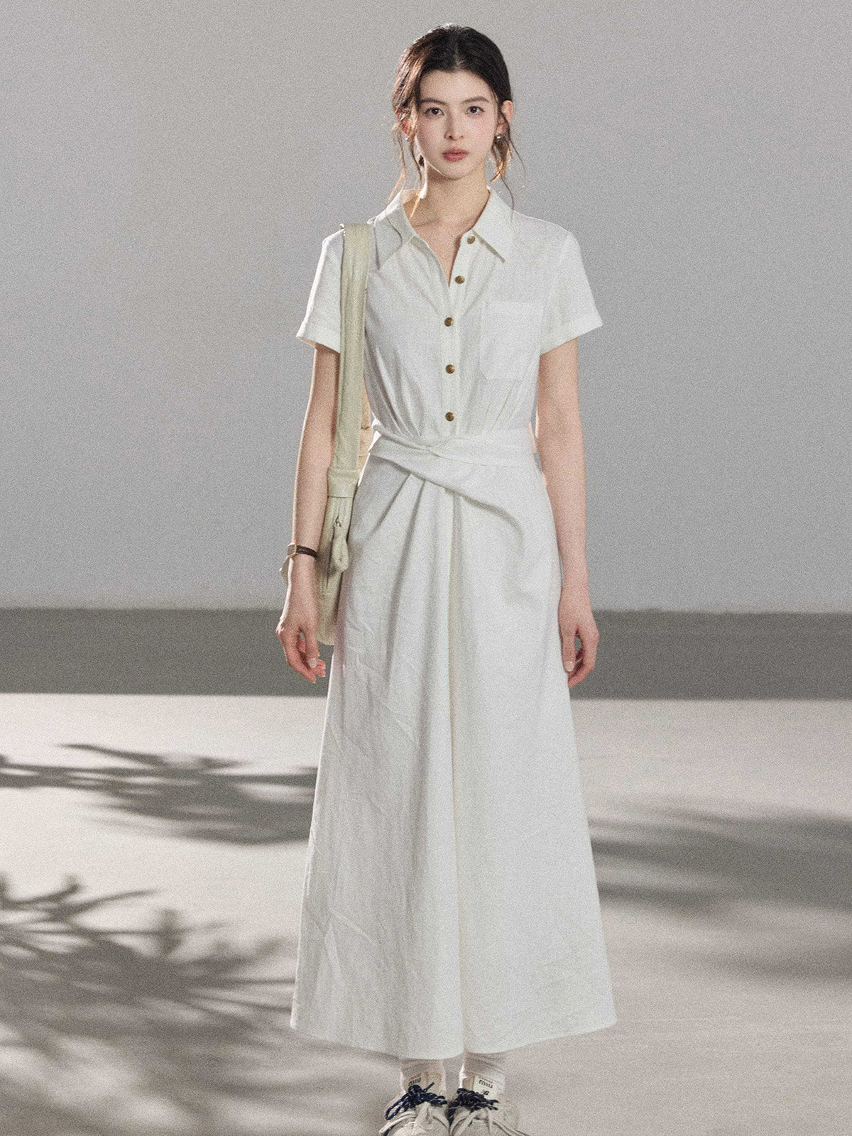 A woman wearing a white A-line dress with a button-front, collar, and twisted waist tie.