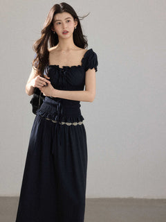 A woman wearing a black long dress with a square neckline, puff sleeves, and a metallic chain belt.