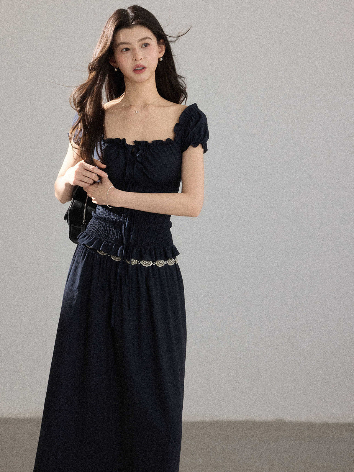 A woman wearing a black long dress with a square neckline, puff sleeves, and a metallic chain belt.