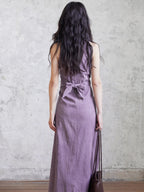 A woman in a purple sleeveless maxi dress seen from behind, showing the back tie belt.
