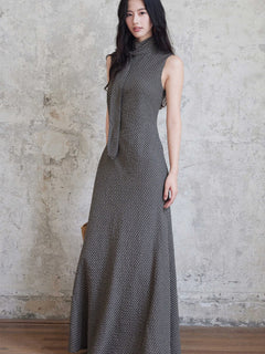 A woman in a sleeveless black gingham floor-length dress with a high neck and tie detail, standing against a textured wall.