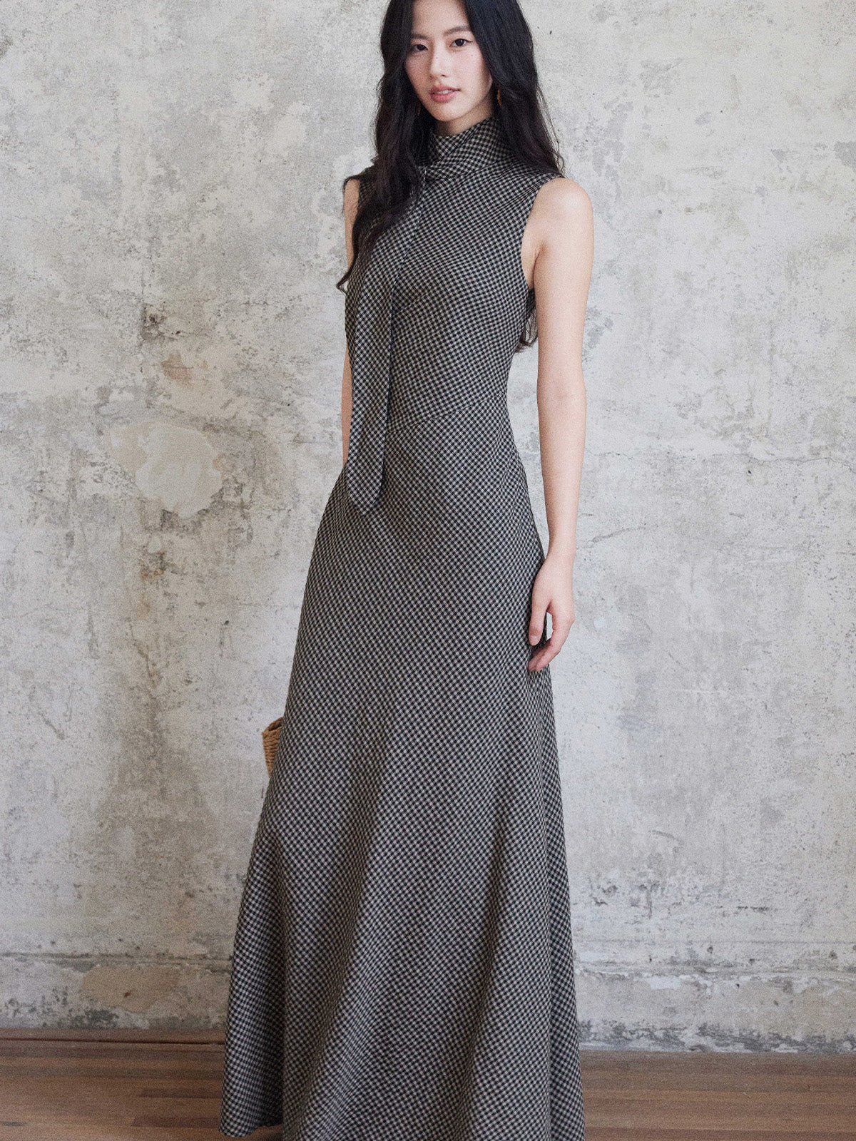 A woman in a sleeveless black gingham floor-length dress with a high neck and tie detail, standing against a textured wall.