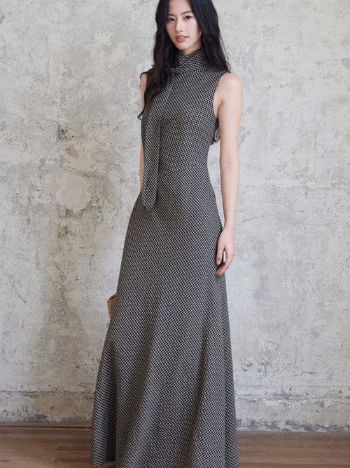 A woman in a sleeveless black gingham floor-length dress with a high neck and tie detail, standing against a textured wall.