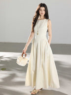 A woman in a cream sleeveless A-line floor-length dress, holding a round handbag with wooden handles.