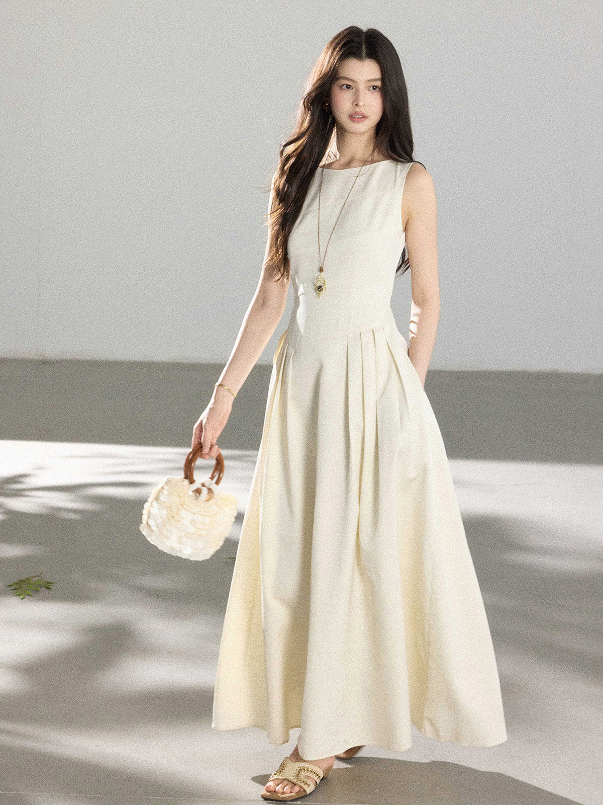 A woman in a cream sleeveless A-line floor-length dress, holding a round handbag with wooden handles.
