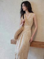 A woman in a beige wrap dress with a V-neck and asymmetrical hemline, leaning against a wall while holding a woven bag.