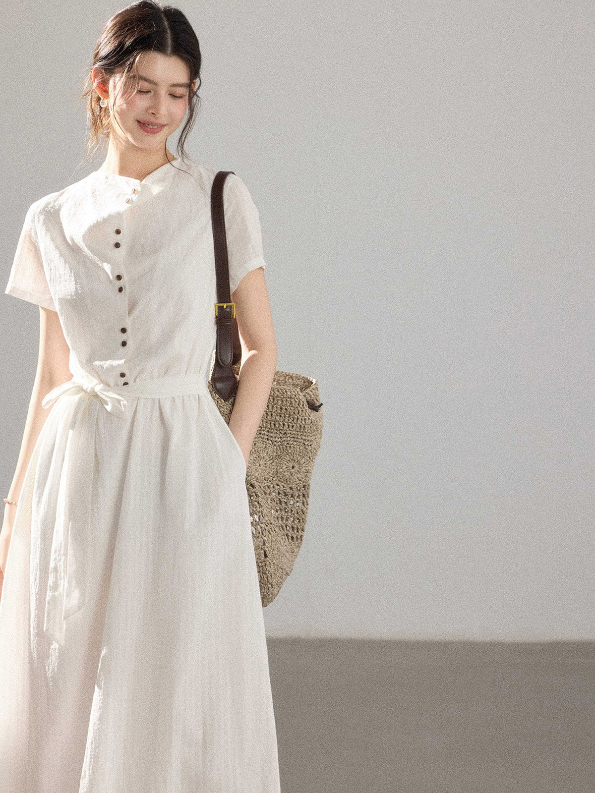 A woman in a white A-line dress with a button placket and tie belt, carrying a woven bag.