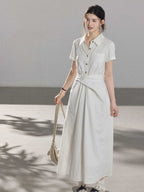 A woman wearing a white A-line dress with a button-front, collar, and twisted waist tie.