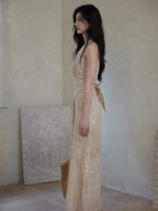 A side profile of a woman in a beige wrap dress with a V-neck and asymmetrical hemline, standing next to a woven bag.